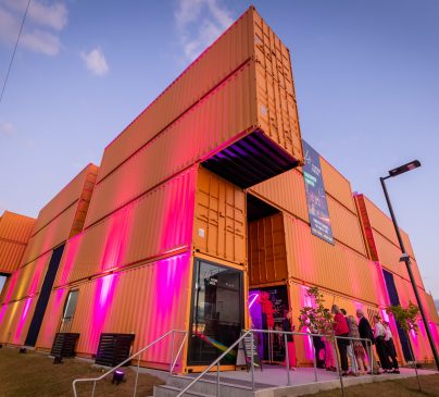 A modern building constructed from stacked shipping containers, illuminated with pink and purple lights. People gather near the entrance.