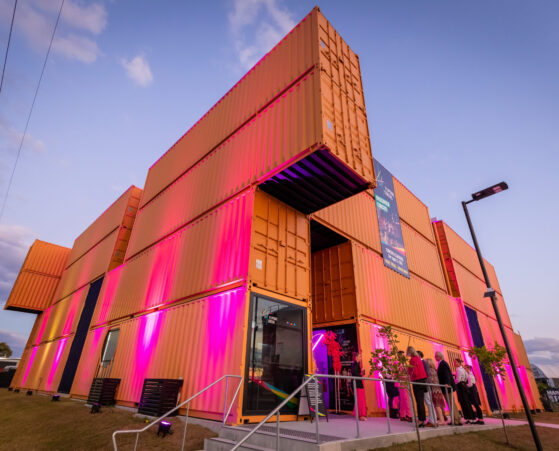 A modern building constructed from stacked shipping containers, illuminated with pink and purple lights. People gather near the entrance.