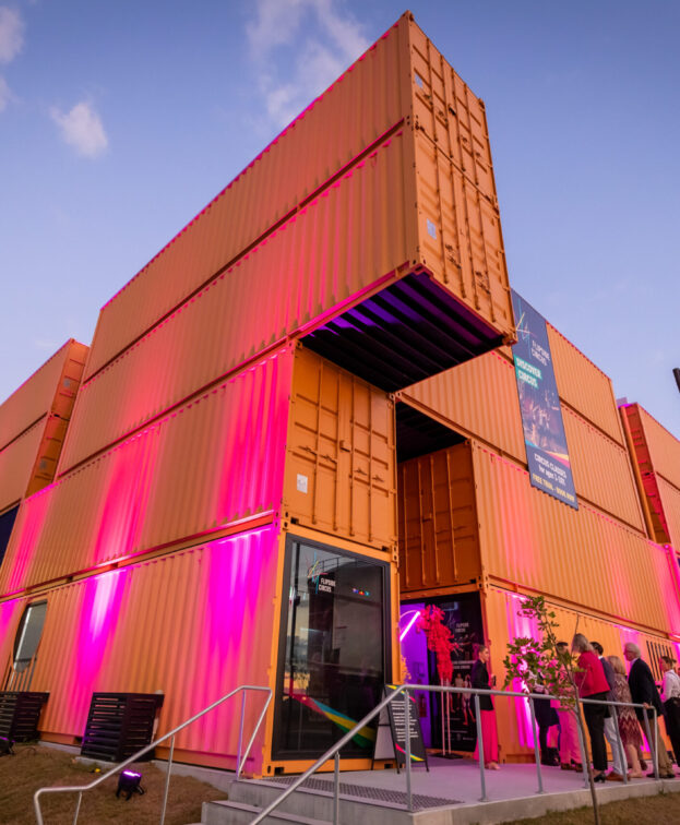 A modern building constructed from stacked shipping containers, illuminated with pink and purple lights. People gather near the entrance.