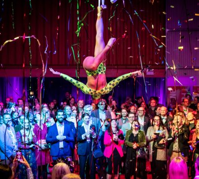 An acrobat in a green outfit performs an aerial act above a crowd at an indoor event at Flipside Circus. Colourful confetti and streamers fill the air.