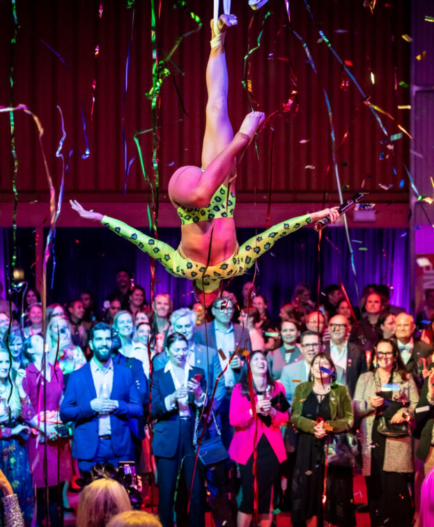 An acrobat in a green outfit performs an aerial act above a crowd at an indoor event at Flipside Circus. Colourful confetti and streamers fill the air.