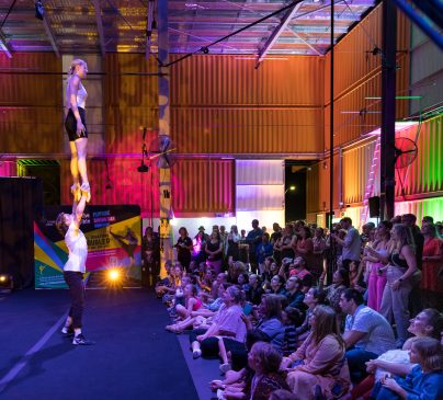 A crowd watches two acrobats perform an impressive hand balance routine inside Flipside Circus. The audience, seated on the floor and standing, observes attentively, surrounded by vivid lights and industrial decor.