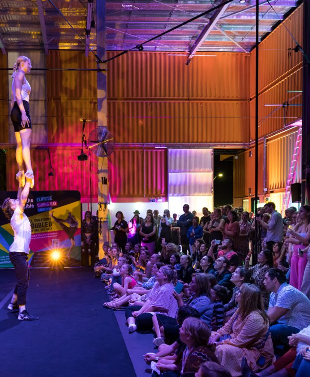 A crowd watches two acrobats perform an impressive hand balance routine inside Flipside Circus. The audience, seated on the floor and standing, observes attentively, surrounded by vivid lights and industrial decor.