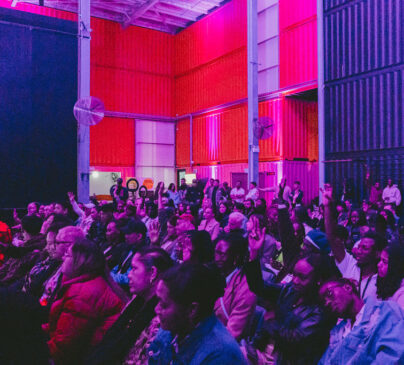 A diverse audience sits in a dimly lit room with vibrant purple and pink lighting. The space has industrial elements like large fans and shipping container walls walls.