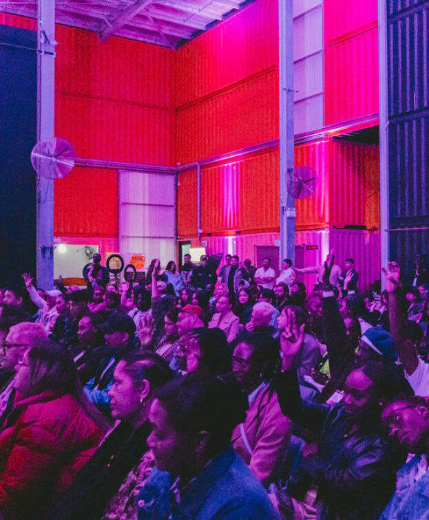 A diverse audience sits in a dimly lit room with vibrant purple and pink lighting. The space has industrial elements like large fans and shipping container walls walls.